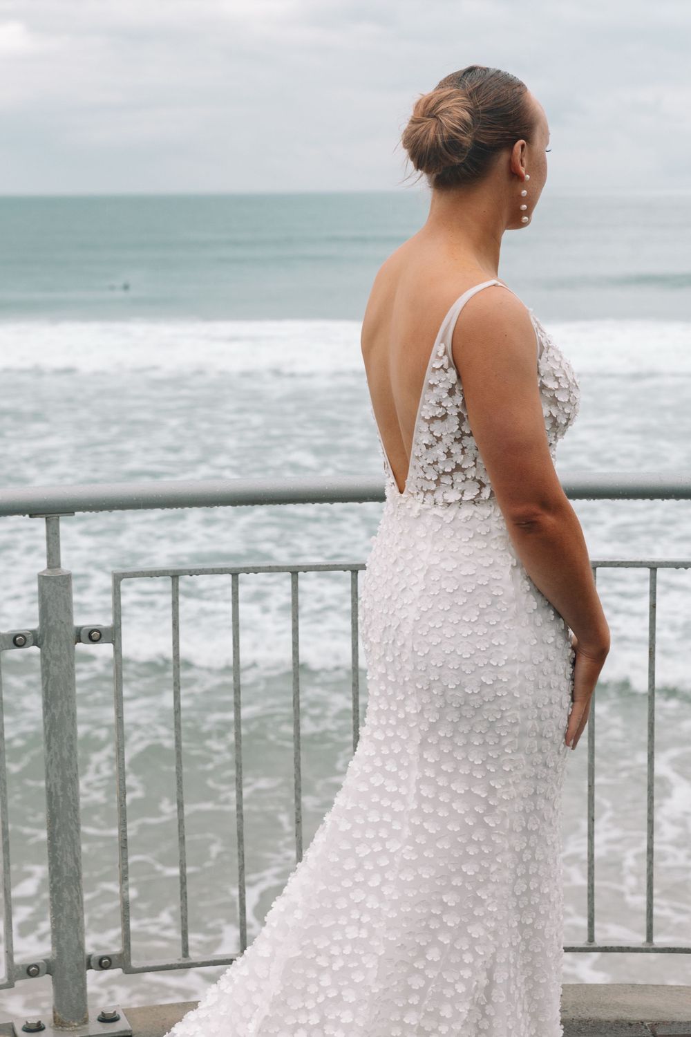 Back view of a bride in a detailed white lace wedding dress gazing at the ocean.
