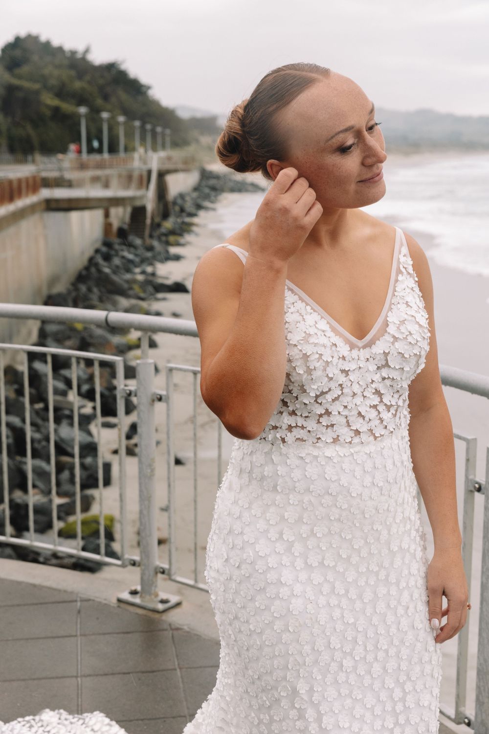 Bride in intricate lace gown adjusts earring while standing near coastal railing.