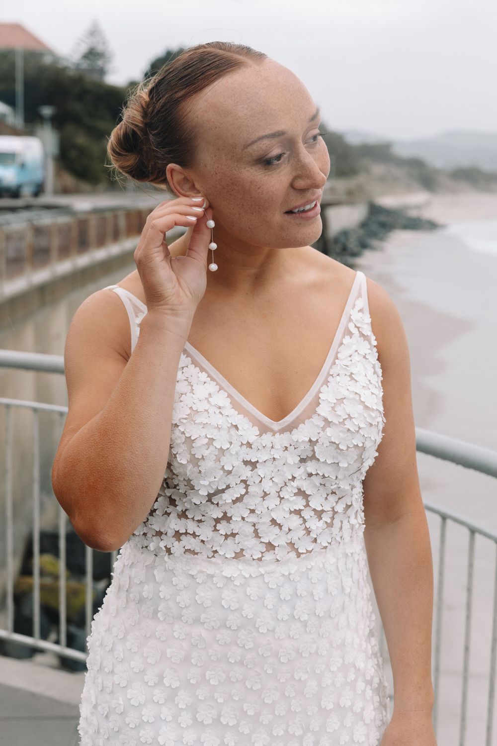A bride in white lace dress with pearl earrings poses against coastal scenery.