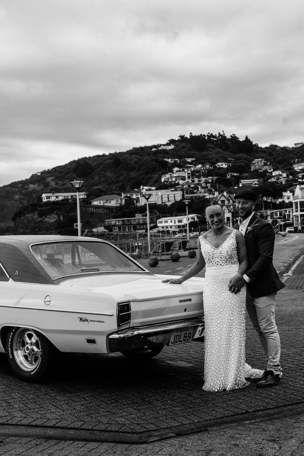 A vintage white car against a backdrop of coastal hillside homes on a cloudy day.