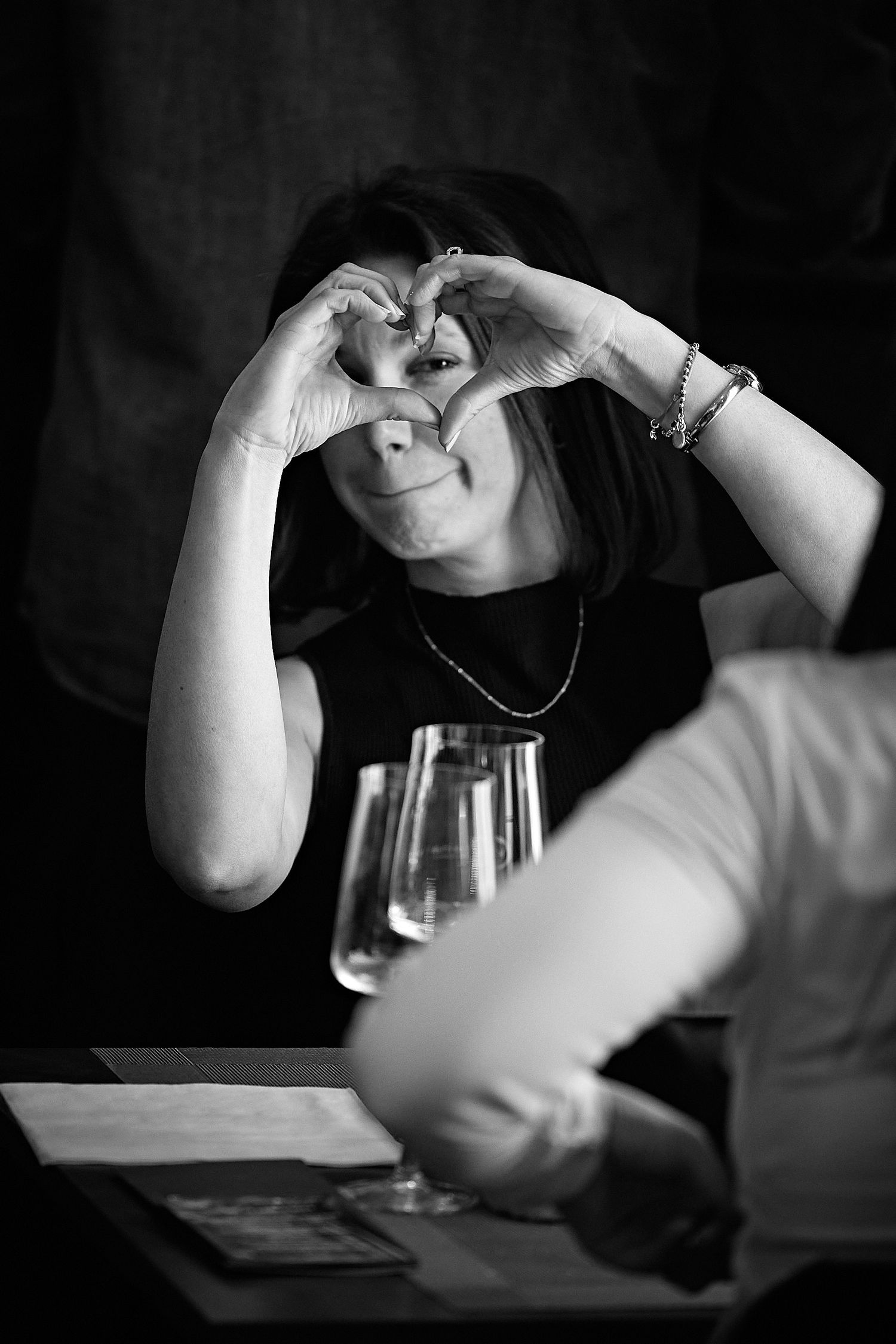 Someone making a heart shape with their hands while seated at a table in black and white photography.