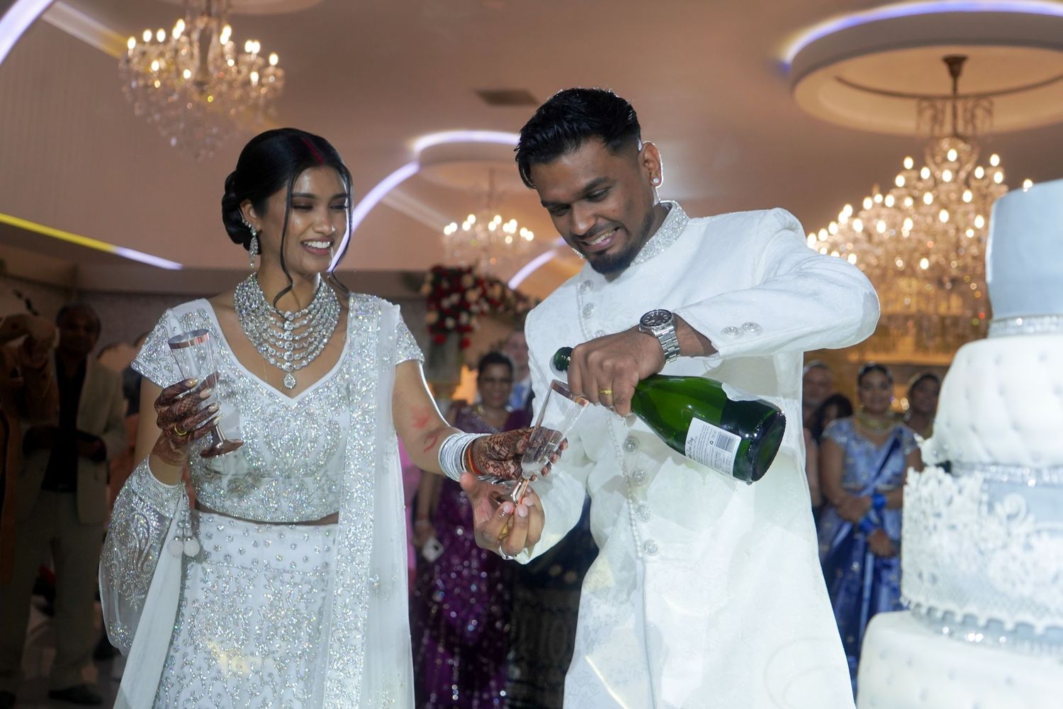 Couple celebrating at their wedding reception while pouring champagne near an elegant white tiered cake.