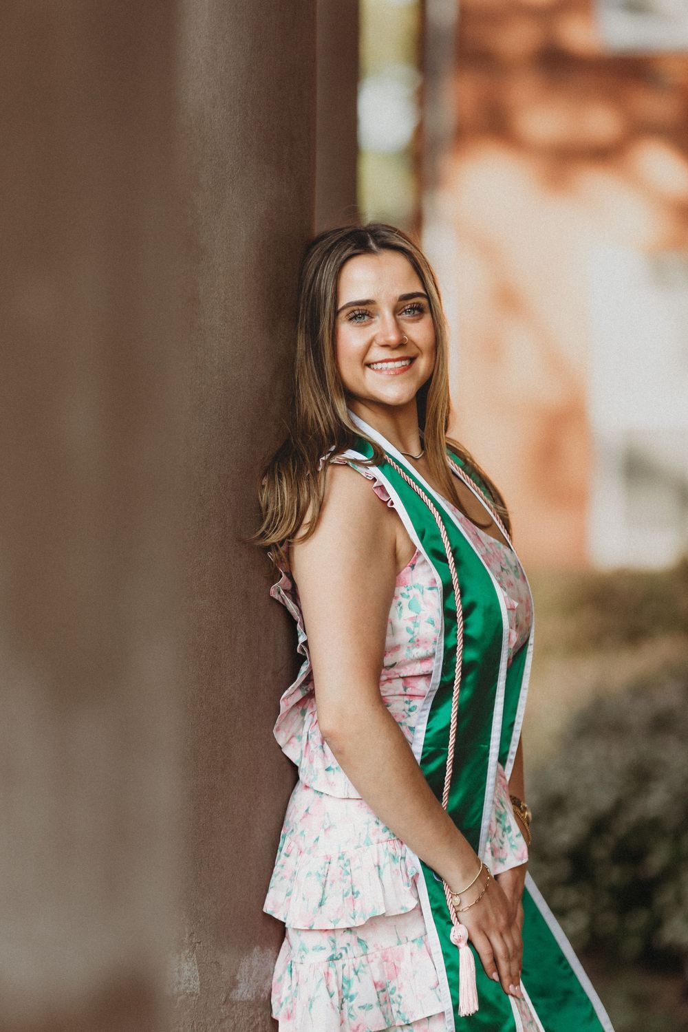 Smiling graduate wearing a pink floral dress with ruffles and green sash poses during outdoor portrait session.