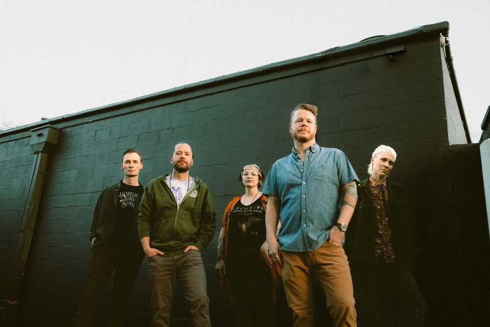 A group of five people stand together in casual attire against a dark wall in dramatic lighting.
