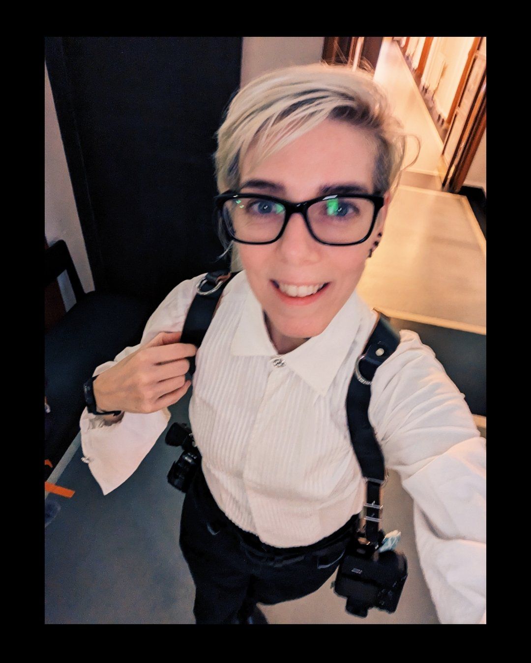 Professional photographer wearing black glasses and white shirt with camera harness taking a selfie.