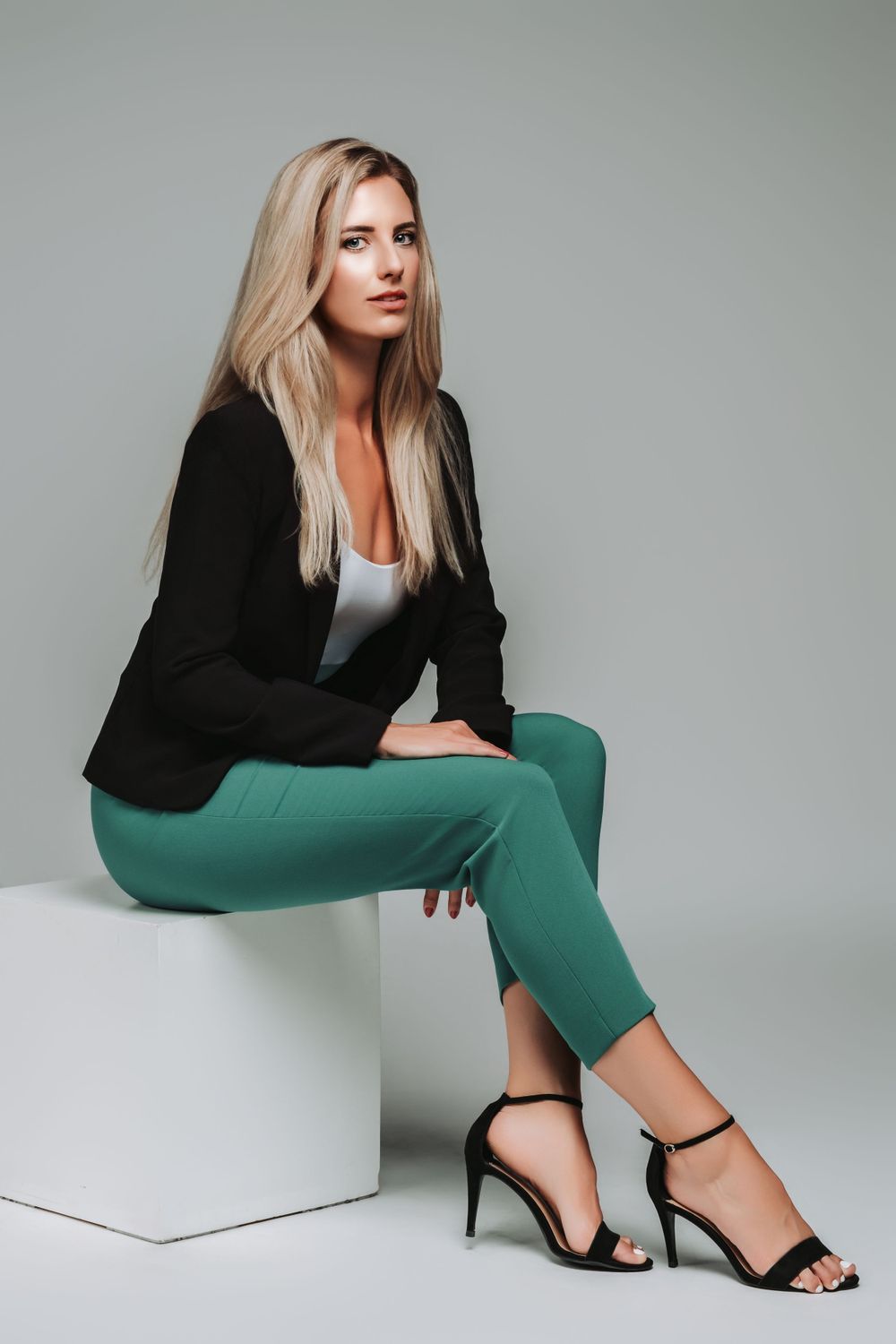 Professional fashion model wearing black blazer, green pants and strappy heels while sitting on white block.