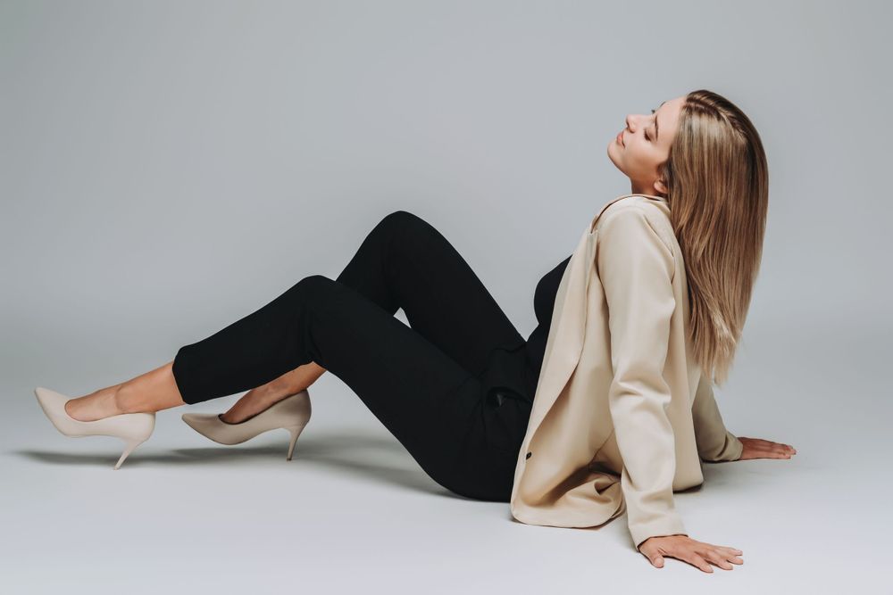 Model in casual outfit lying on floor wearing beige blazer and black pants with neutral heels.