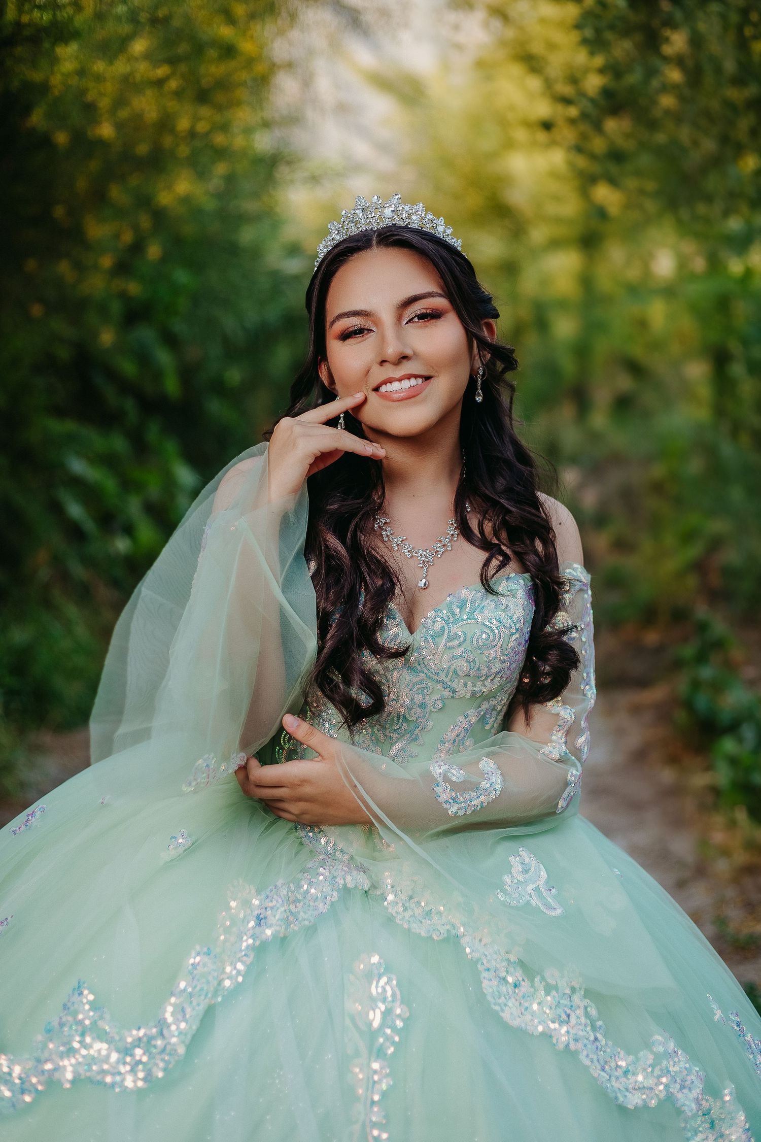 A young woman in a mint green quinceañera dress with lace details poses outdoors in a natural setting.