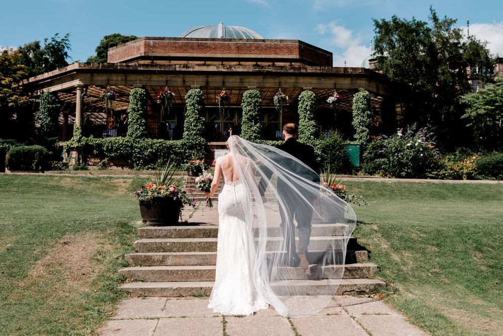 Bride with flowing veil stands on stone steps leading to garden pavilion on sunny day.