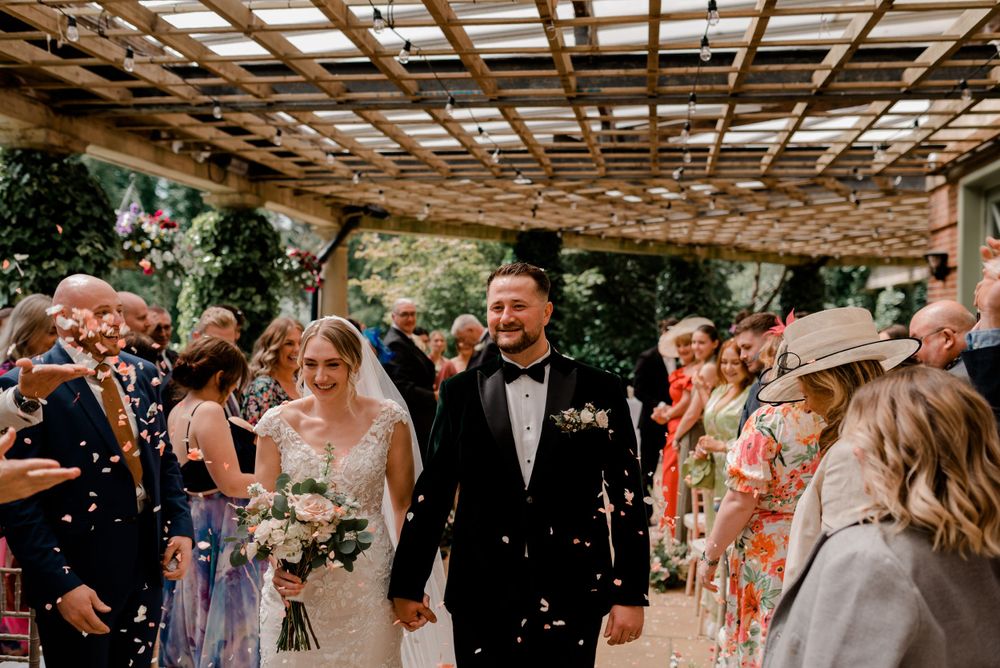 Newlyweds walk down an aisle under a wooden pergola while guests shower them with confetti at an outdoor celebration.