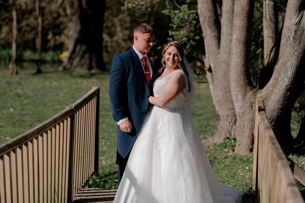 A bride in a white ball gown and groom in a navy suit share a moment together on a wooden bridge in a park setting.