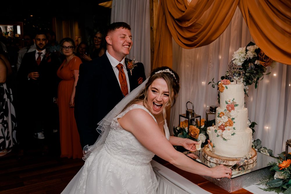 A bride in a white lace dress reaches for a tiered wedding cake at a reception with elegant gold drapery.