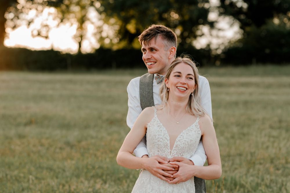 A newlywed couple shares a candid moment together in a sunlit field during golden hour.