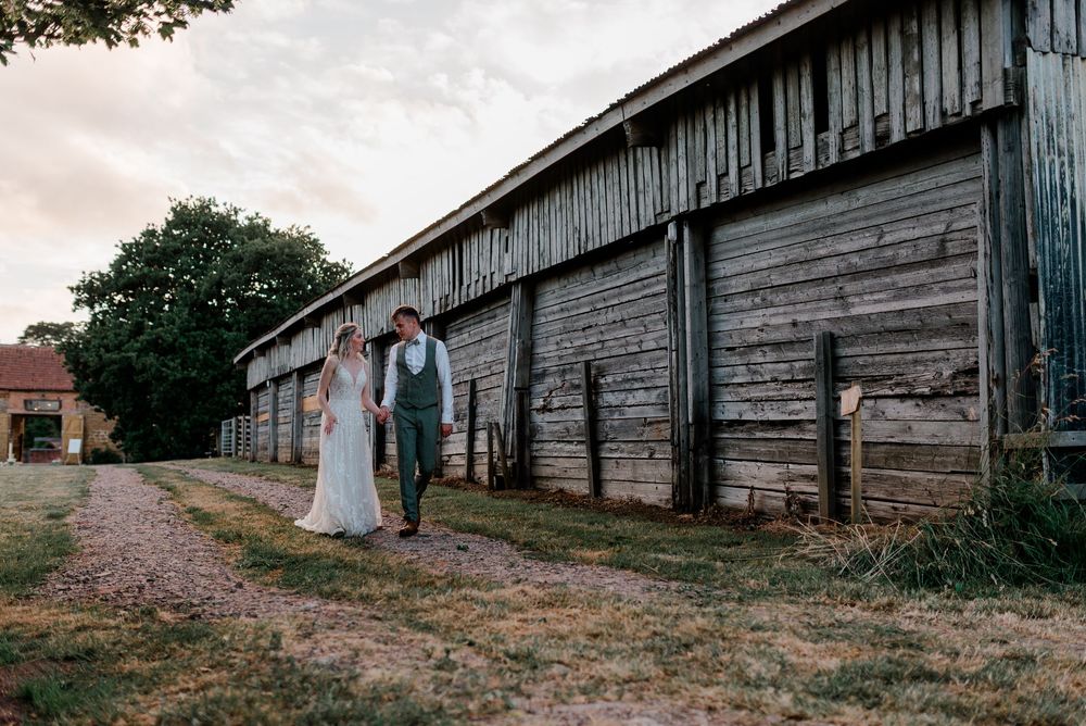 A couple stands by a rustic wooden barn on a dirt path at sunset during their country wedding.