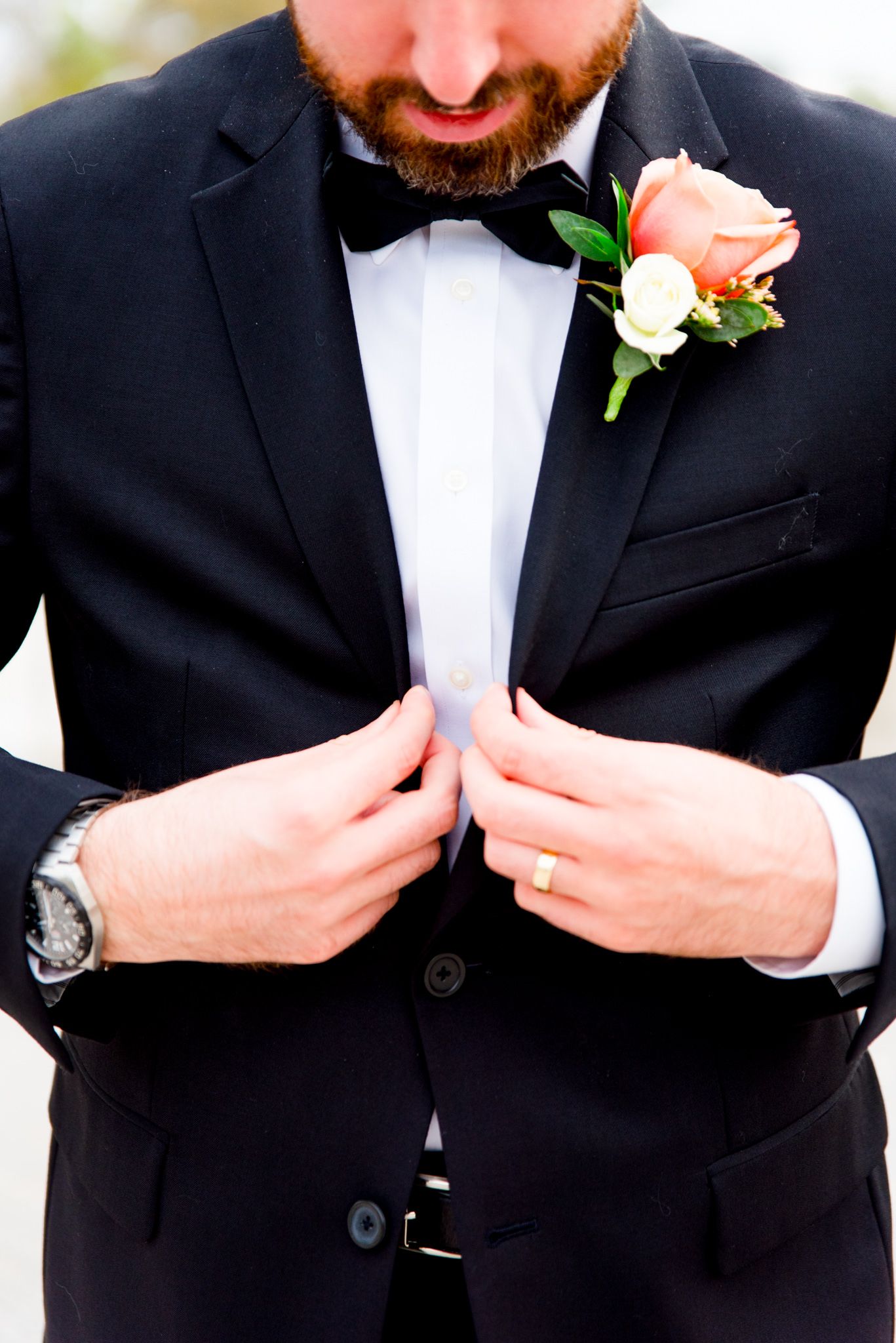 groom with beard buttoning his black suit, showing coral rose boutonniere