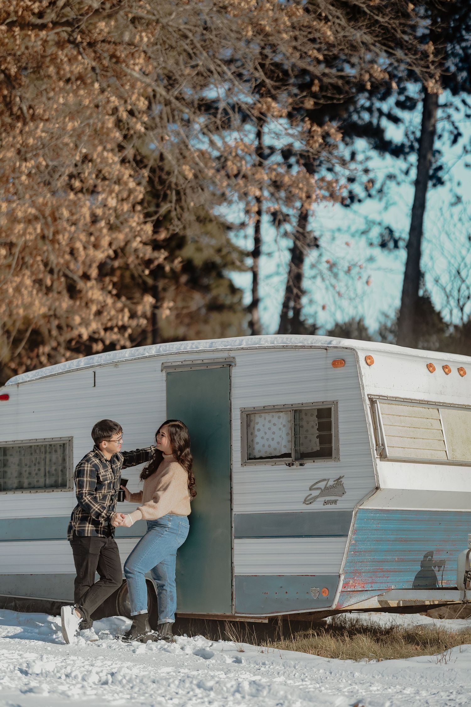 A couple enjoys a winter moment outside their vintage white travel trailer in the snow.