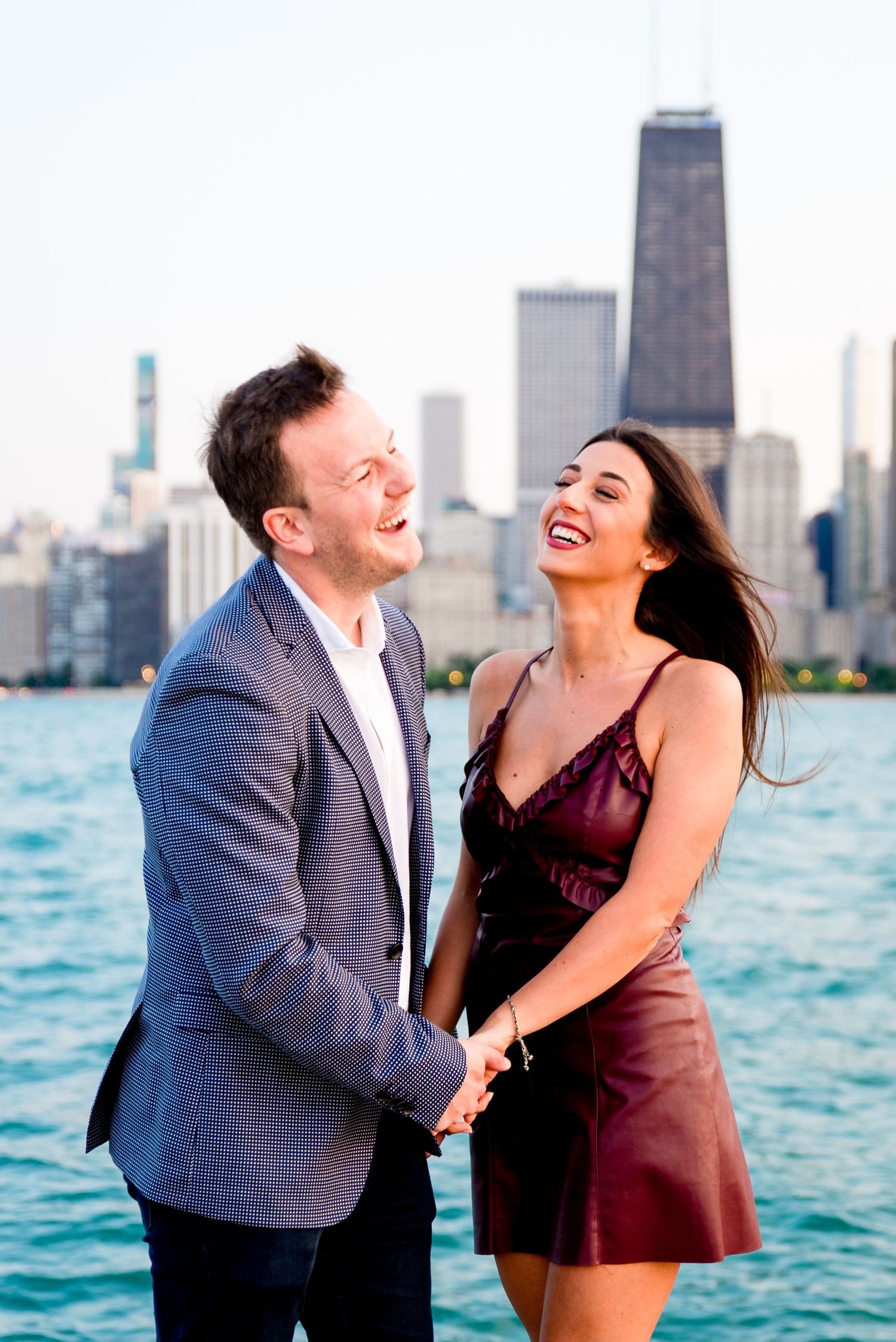 man and woman in formalwear holding hands laughing in front of Chicago skyline after man proposed at North Ave Beach
