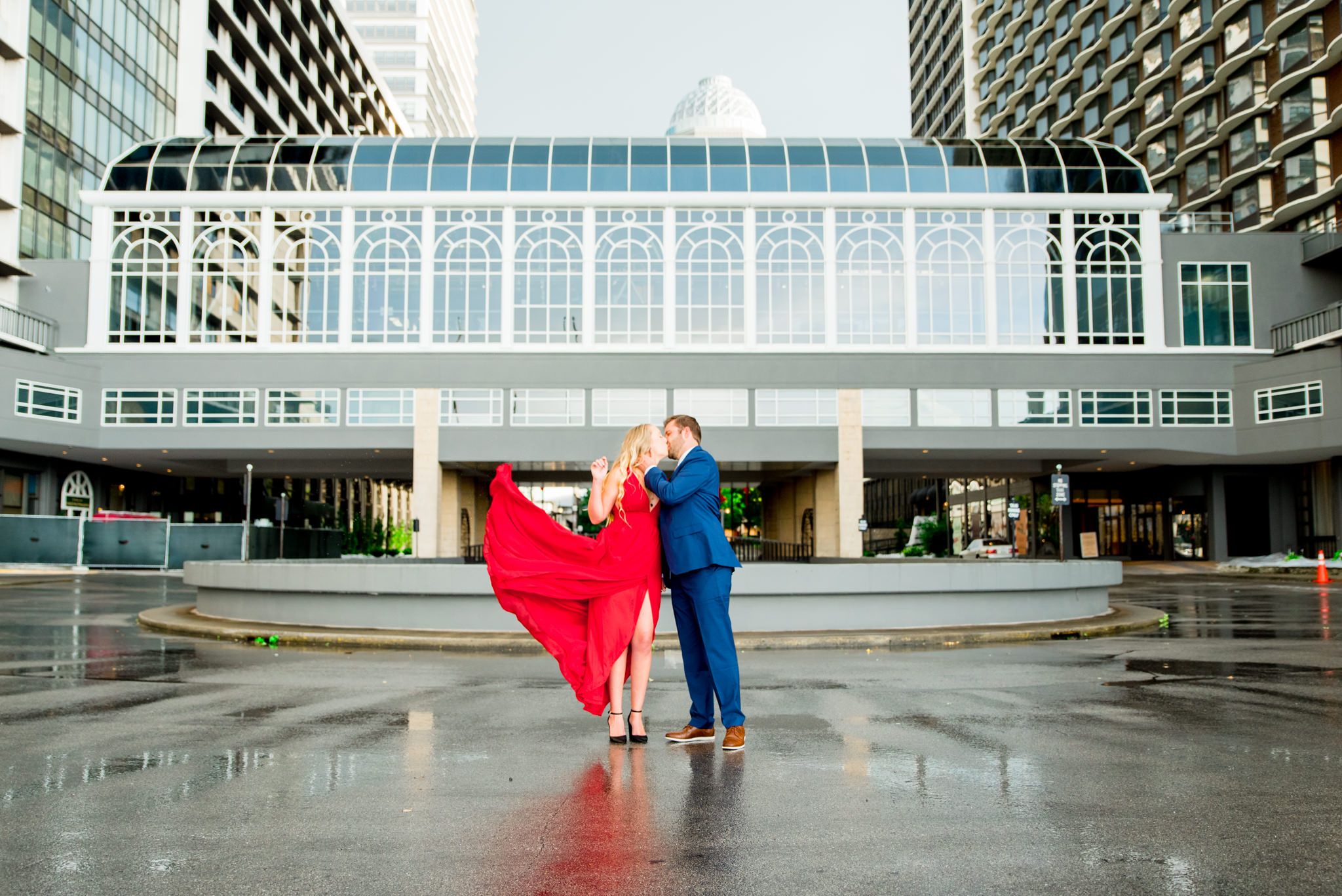 woman in long red dress fans it out in front of Galt House Hotel while fiance in navy suit kisses her