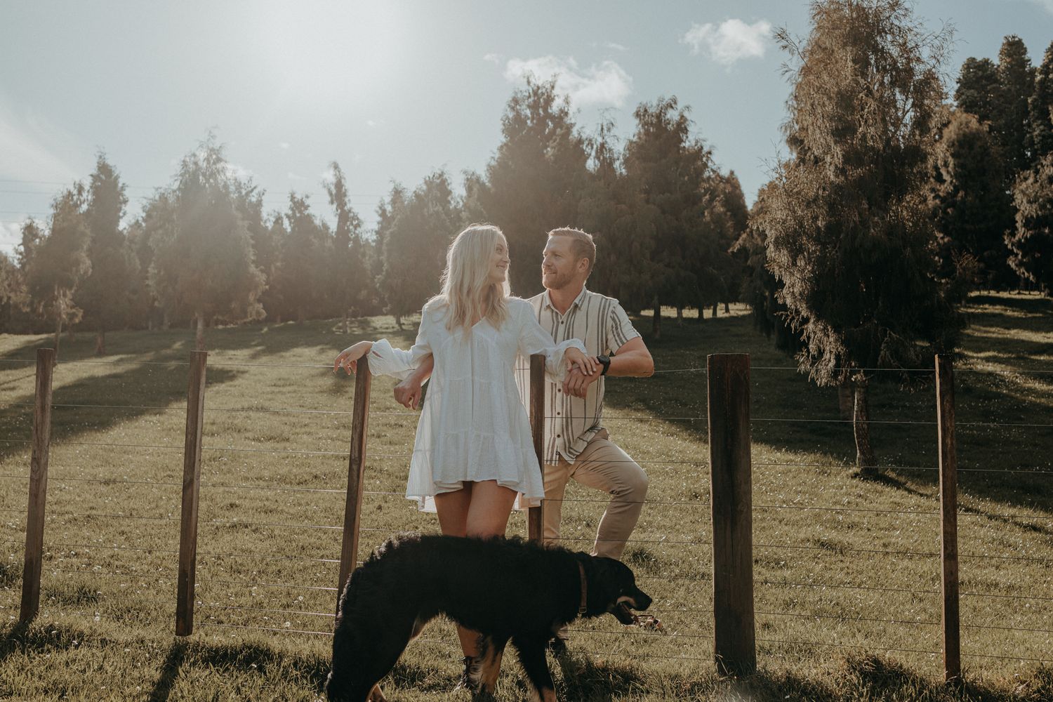 Couple shares a tender moment in a sunny field while their black dog stands nearby.