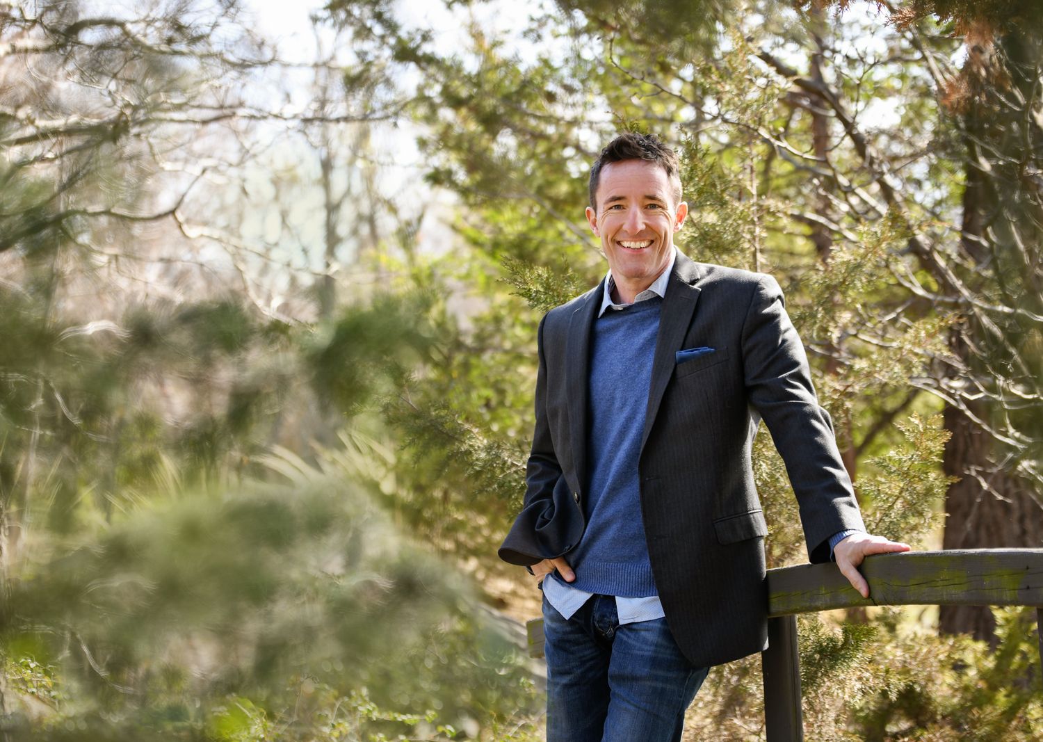 Casual business portrait of someone in a blazer and jeans outdoors among trees.
