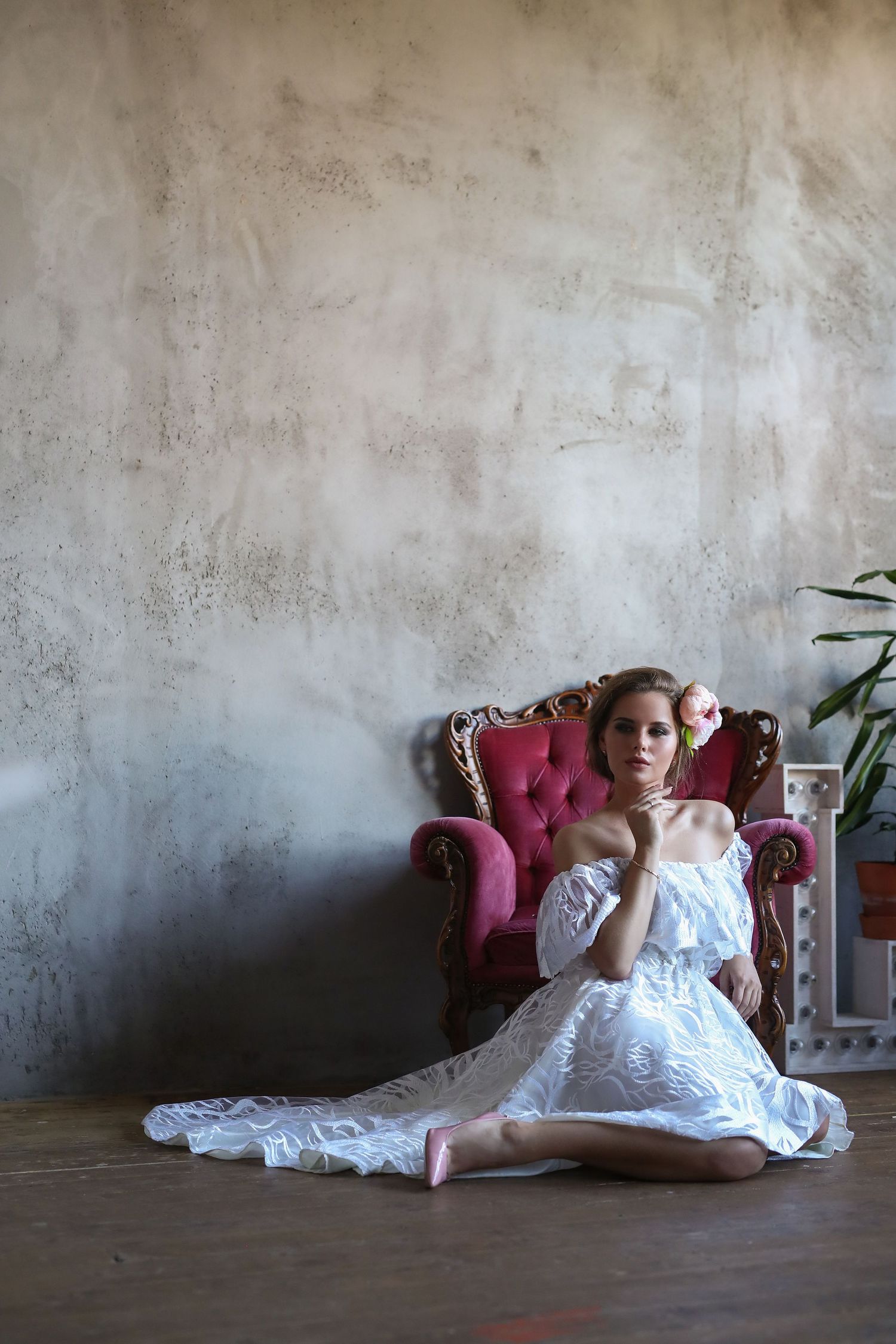 Woman in white lace dress poses elegantly on vintage red velvet chair against textured wall.