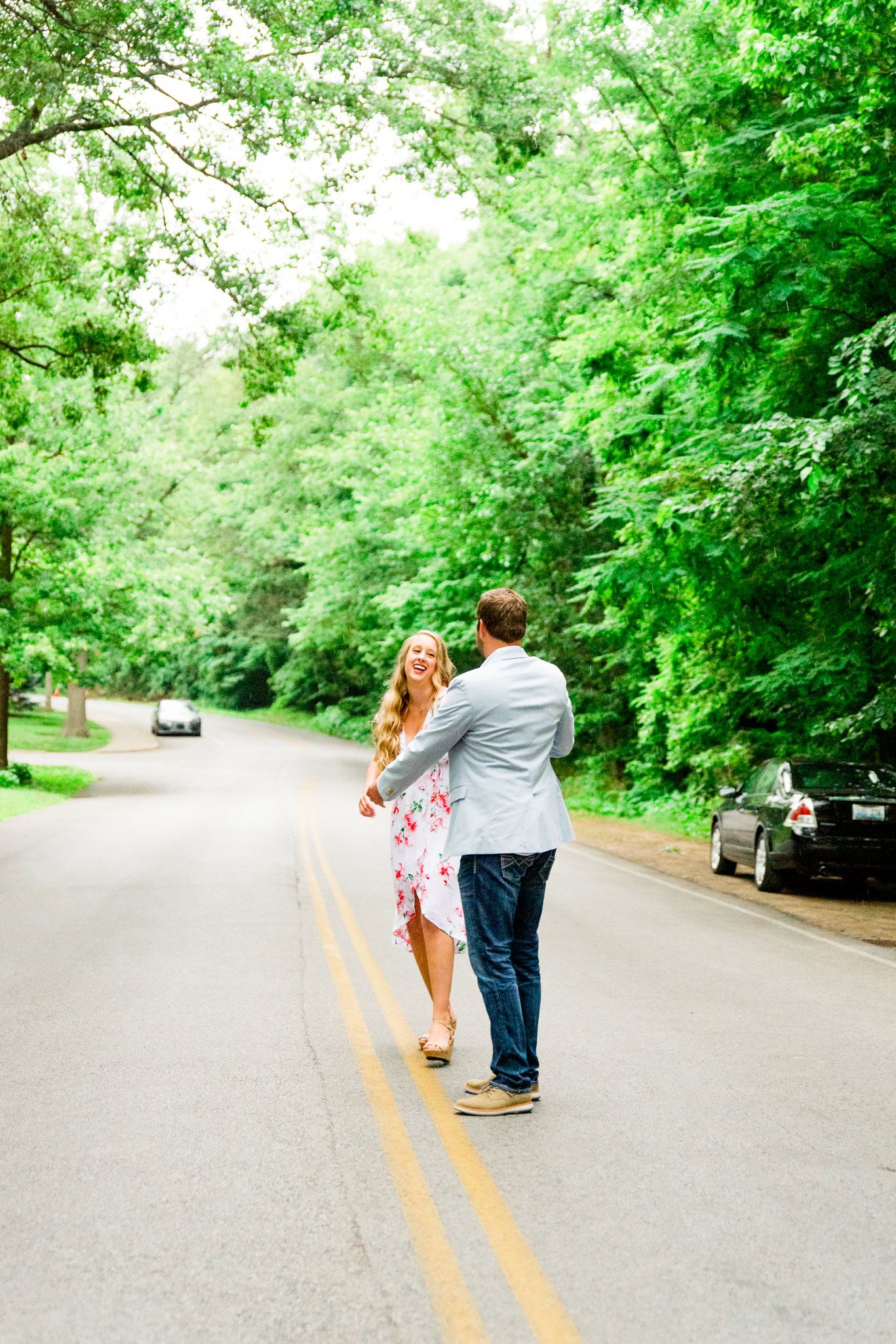engaged couples in floral dress and light blue suit jacket and jeans dancing in middle of road under trees