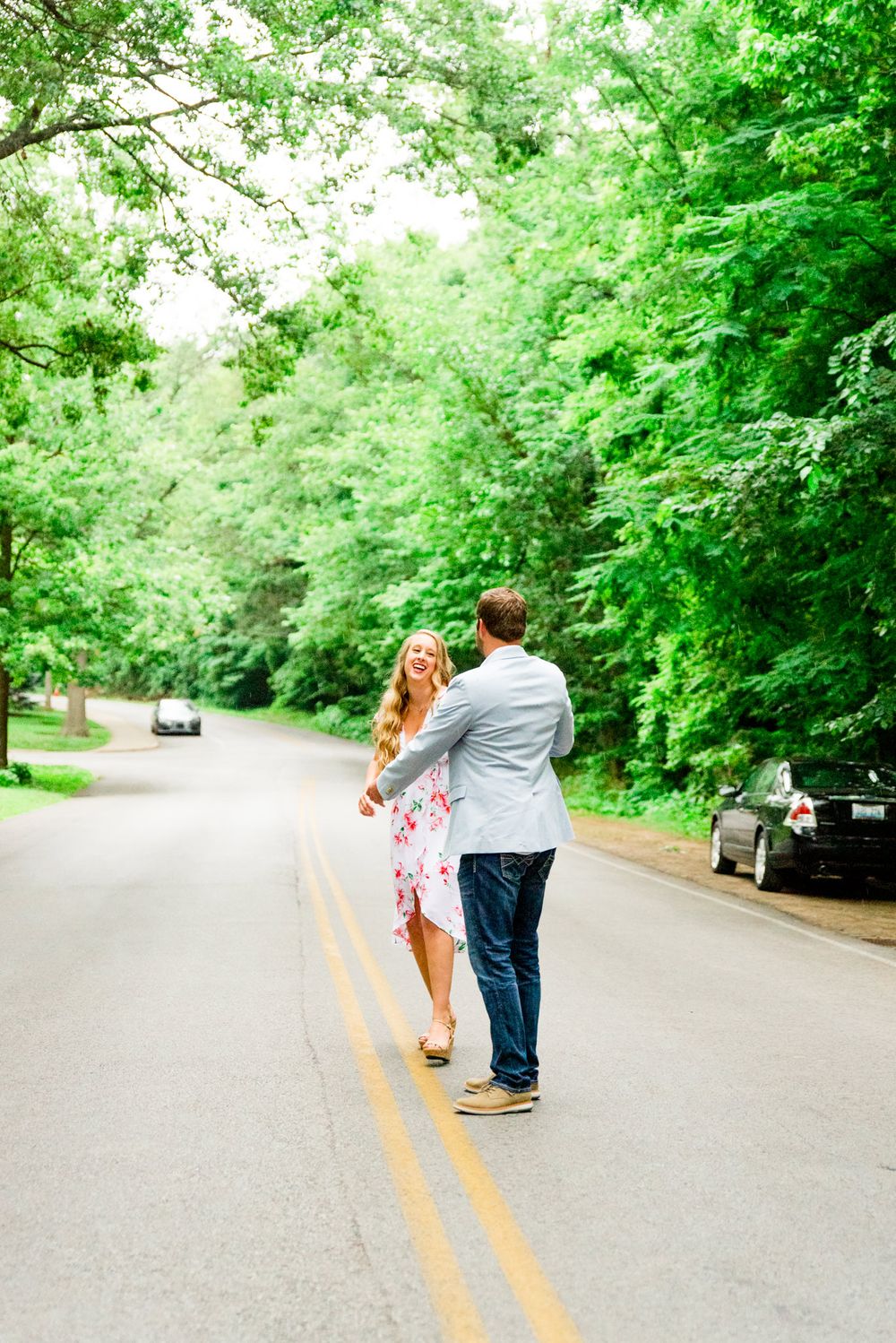 engaged couples in floral dress and light blue suit jacket and jeans dancing in middle of road under trees