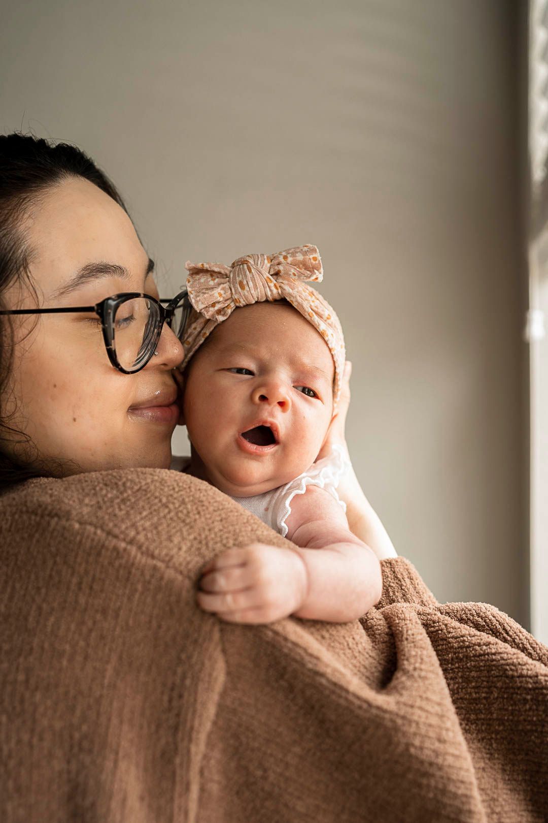 Newborn baby girl yawning in her moms arms. wearing a pink bow. Mom is smiling into her cheek, wearing glasses.