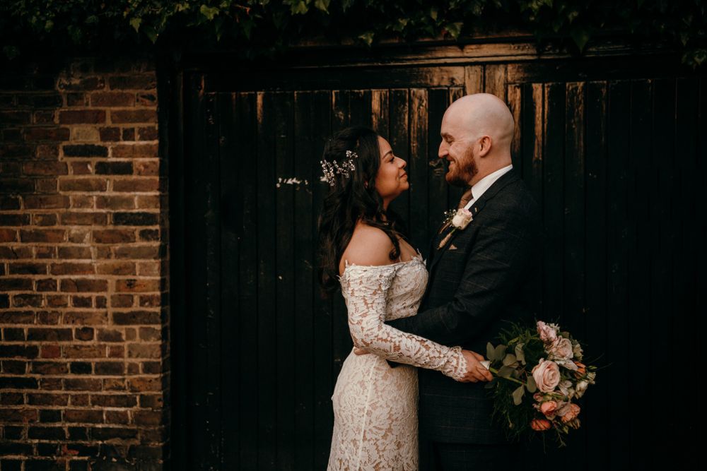 A romantic wedding couple shares an intimate moment against a rustic brick wall while holding a peach floral bouquet.