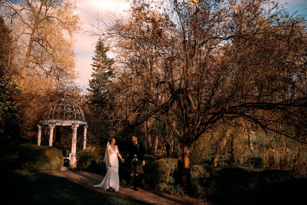 A gazebo stands amidst autumn trees at sunset while a figure in white walks along a garden path.