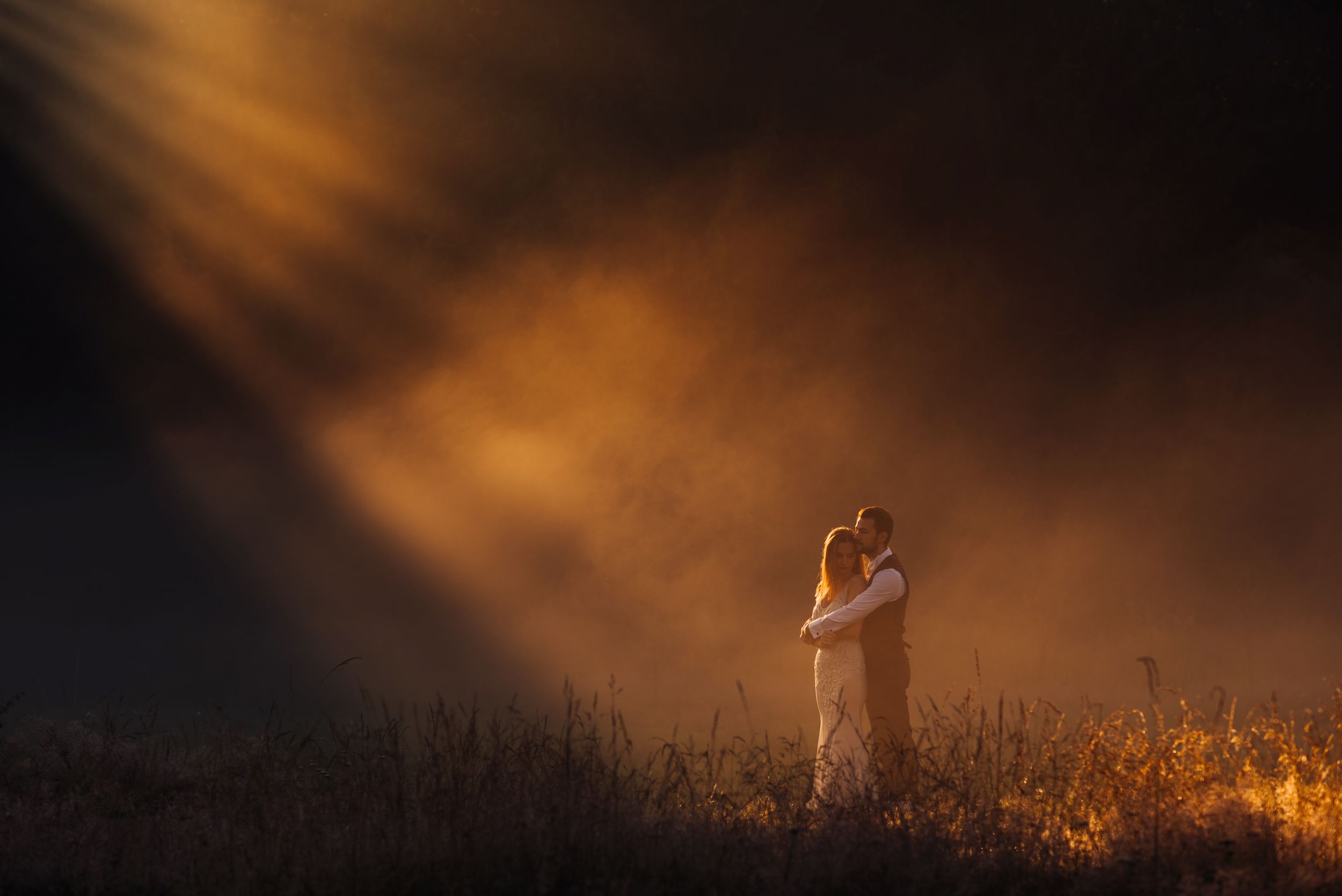 Dramatic silhouette of a couple embracing in a misty field at sunset with warm orange lighting.