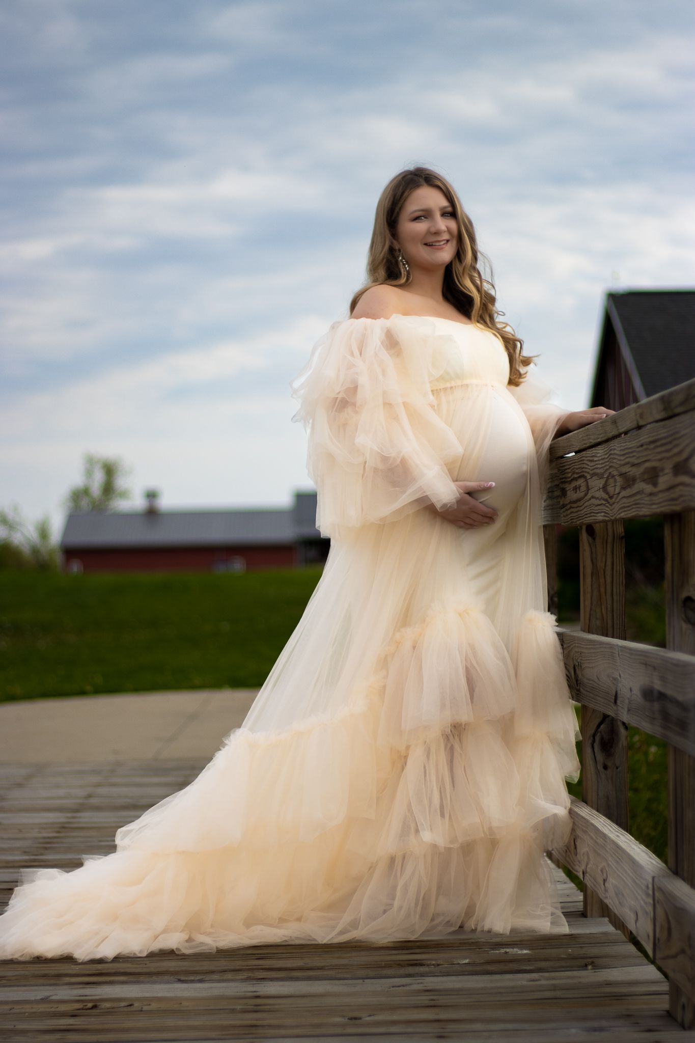 A flowing cream-colored maternity gown photographed on a wooden deck with a barn in the background.