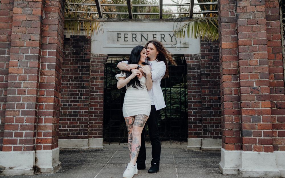 A couple poses together under the Fernery entrance sign on a brick facade.