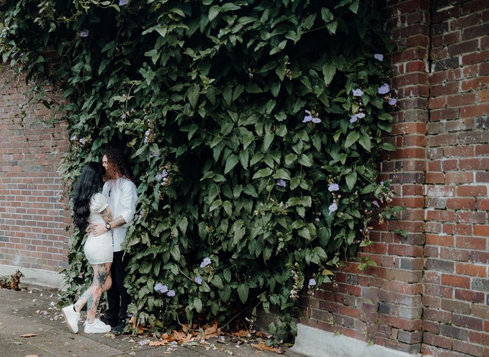 A couple stands against a brick wall covered in climbing green vines.