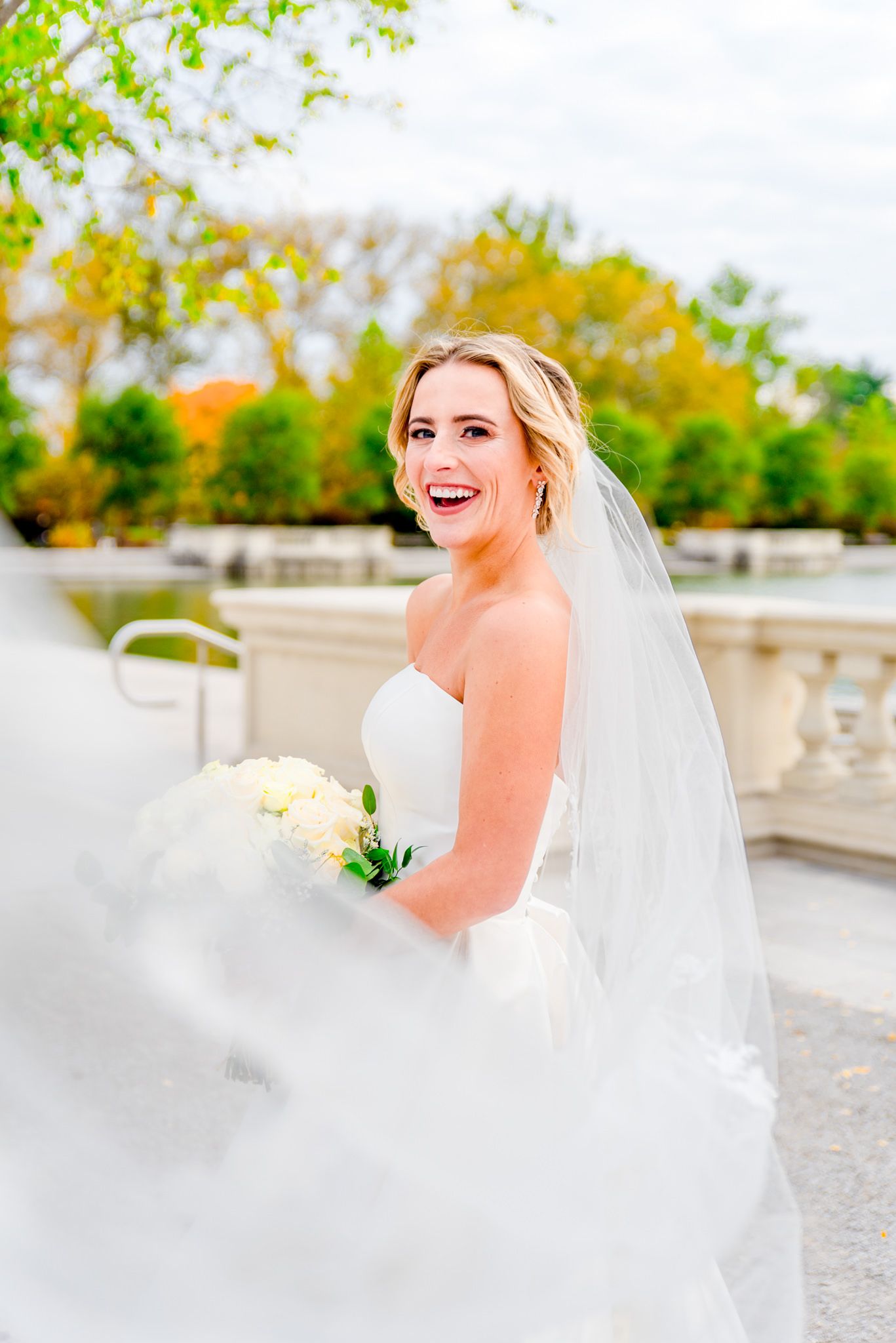 blonde bride with updo and long veil and white bouquet laughing at the camera for Forest Park STL wedding pictures