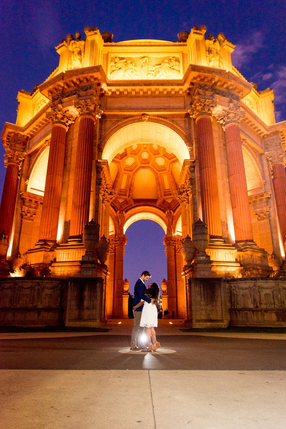 young couple in front of Palace of Fine Arts at night, lit up with blue sky in background
