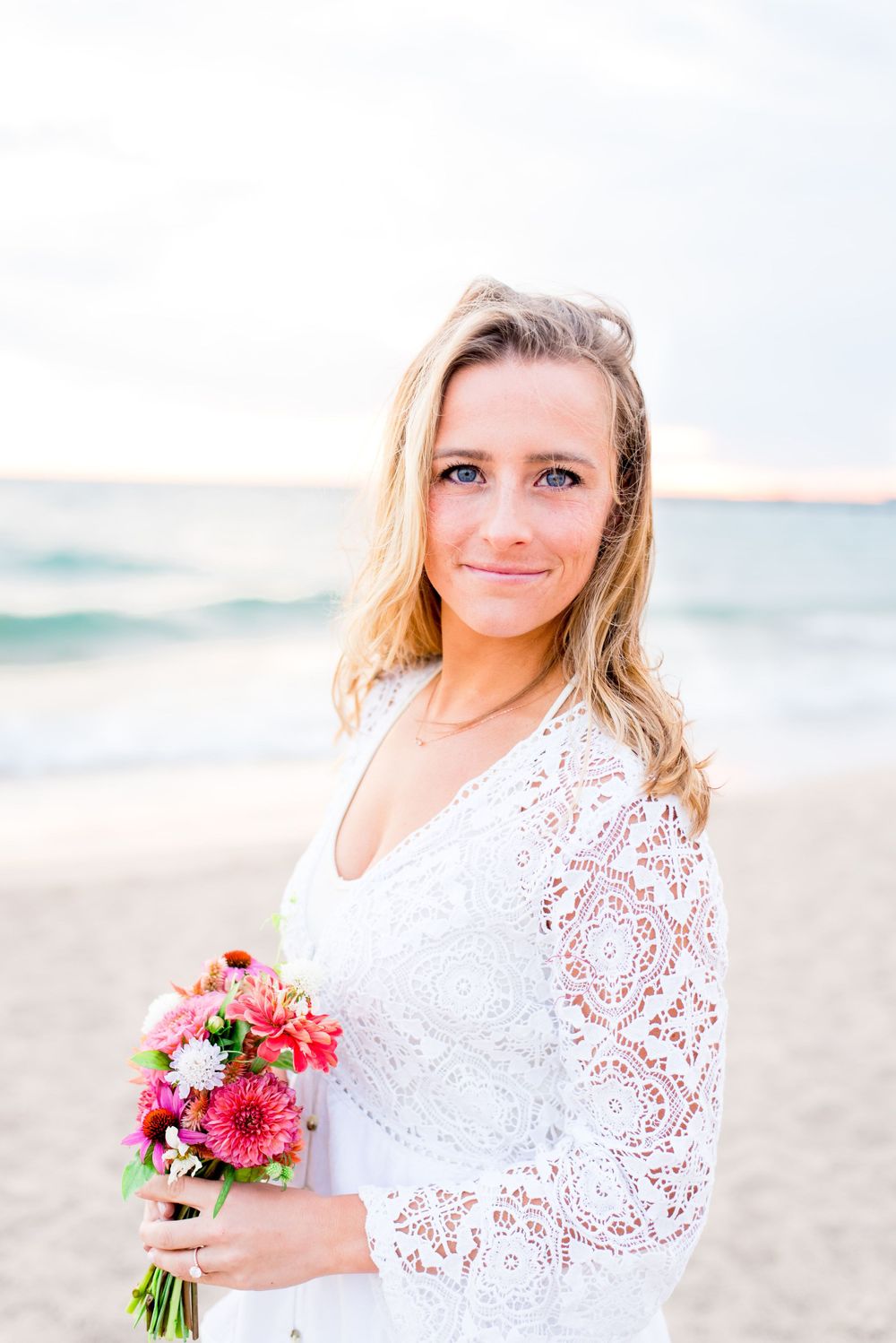 blonde woman in white lace dress softly smiling and holding pink bouquet on North Avenue Beach