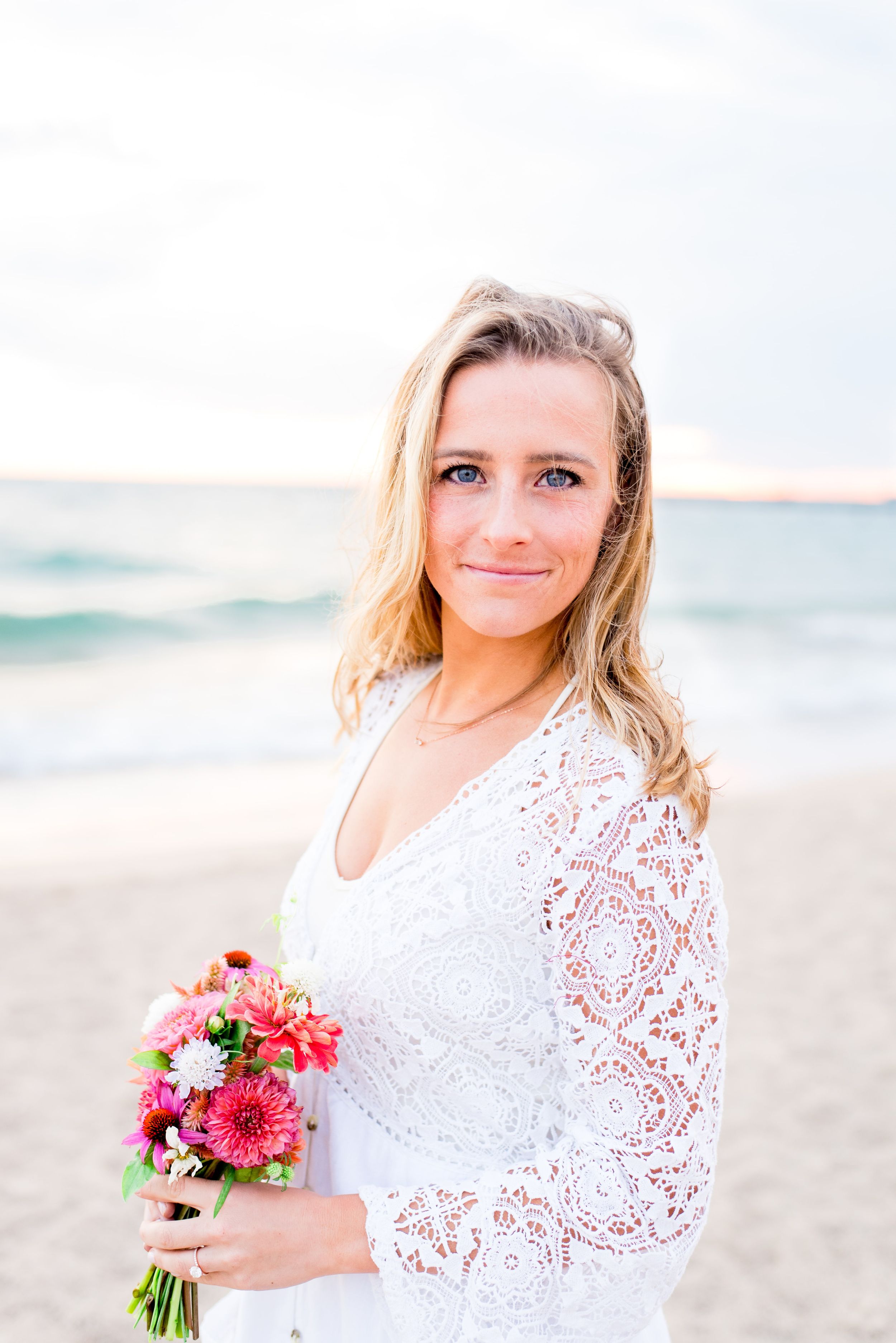 blonde woman in white lace dress softly smiling and holding pink bouquet on North Avenue Beach