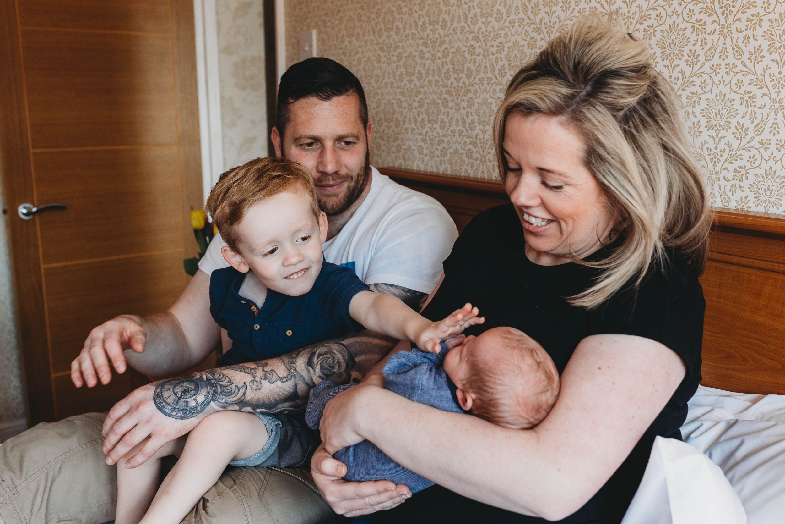toddler boy touching baby boy face sitting with his parents on bed
