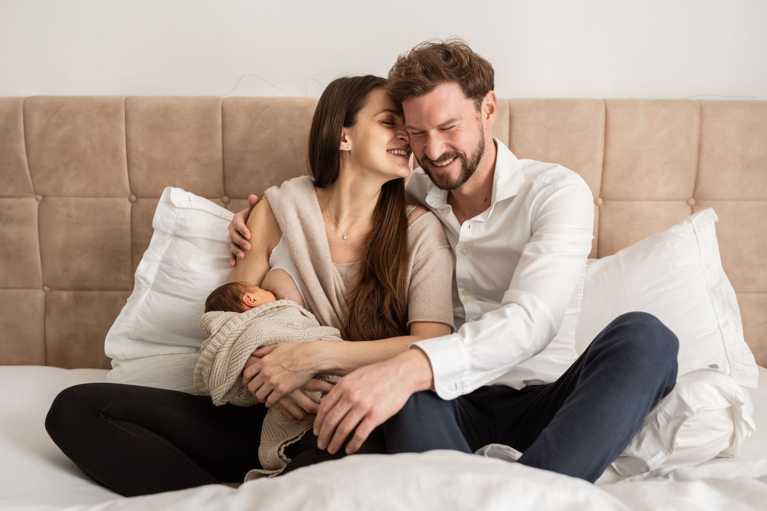 Babyfotograf Stockerau, die Eltern sitzen auf dem Ehebett und kuscheln während die Mama ihr Neugeborenes Baby stillt