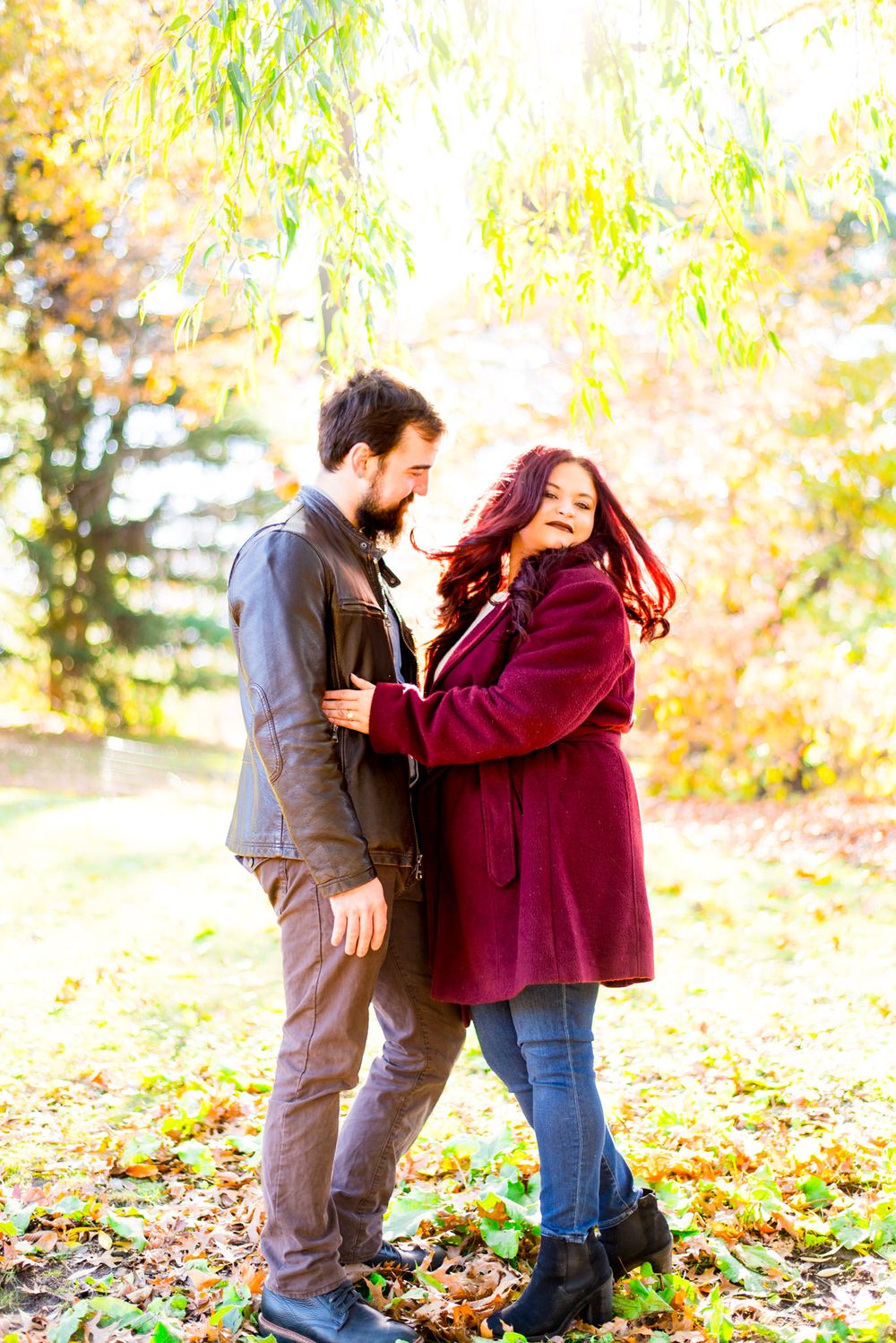 woman in red jacket flips her long red hair under yellow tree at sunrise in Central Park while fiance hugs her