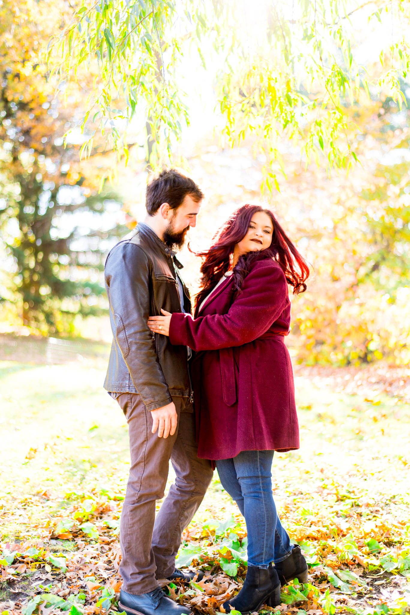 woman in red jacket flips her long red hair under yellow tree at sunrise in Central Park while fiance hugs her