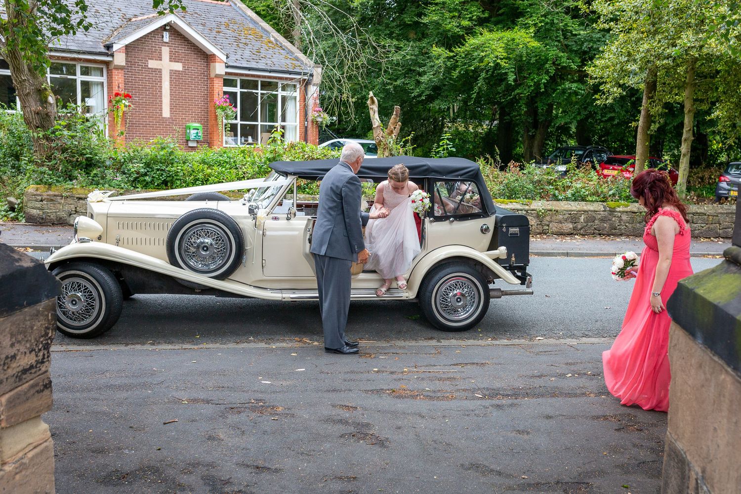 Bride gets help from the chauffeur getting out of the wedding car watched by a bridesmaid