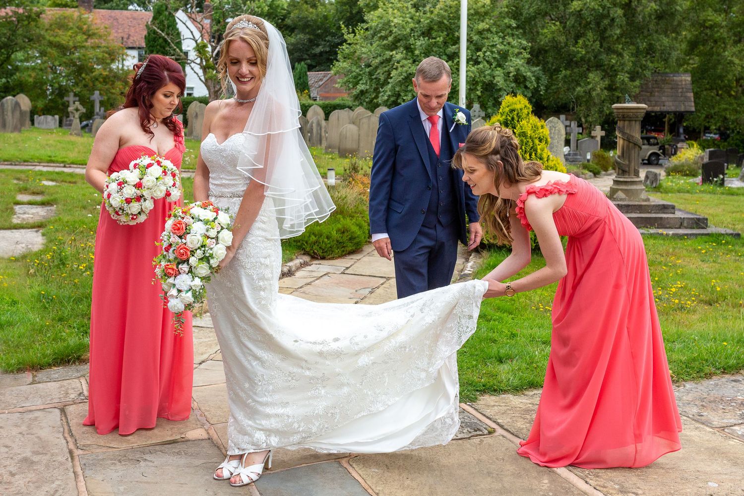 Bridesmaid helps the bride with her dress laying it out watched by her father and another bridesmaid