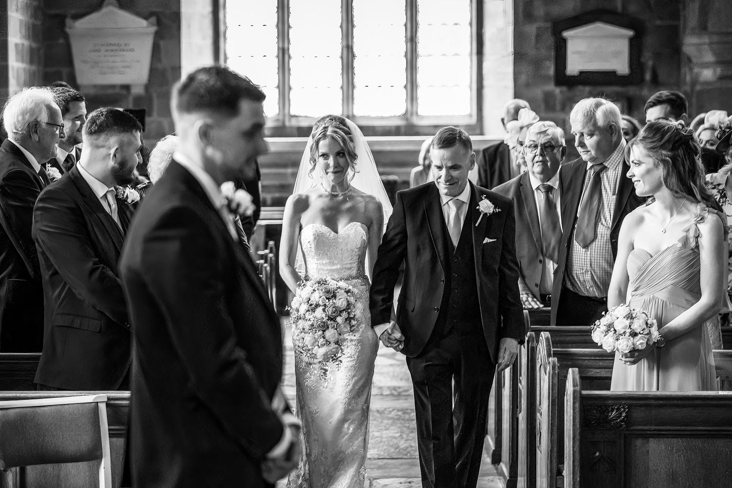 Bride walks down the aisle smiling with her father watched by wedding guests