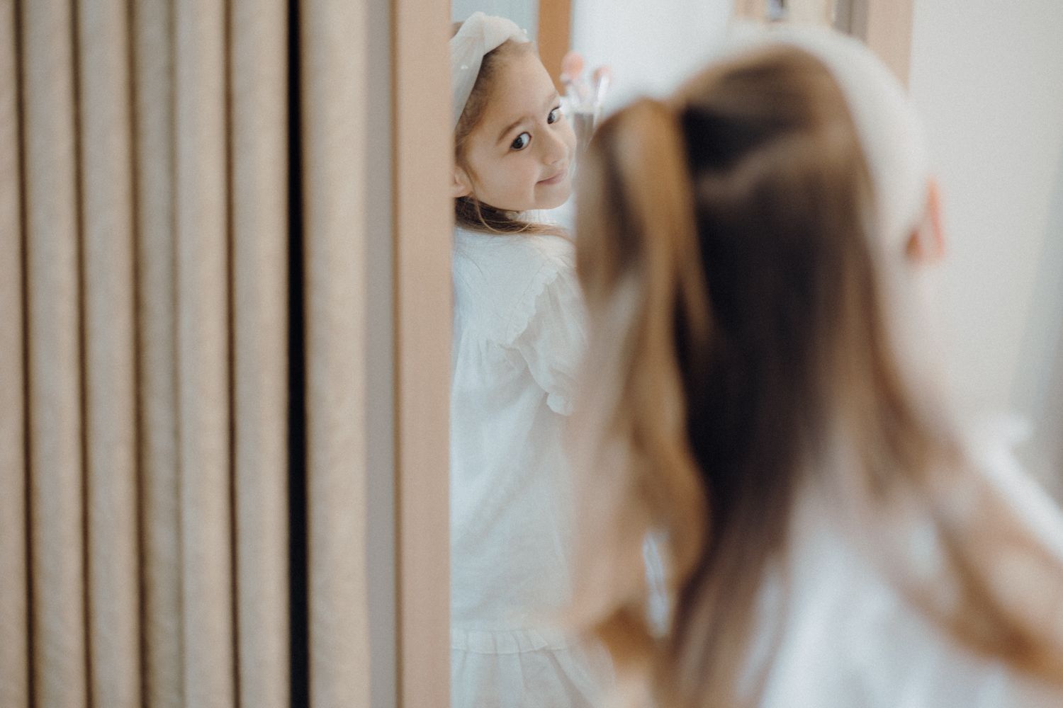 A reflection shot of someone in white clothing looking into a mirror near curtains.