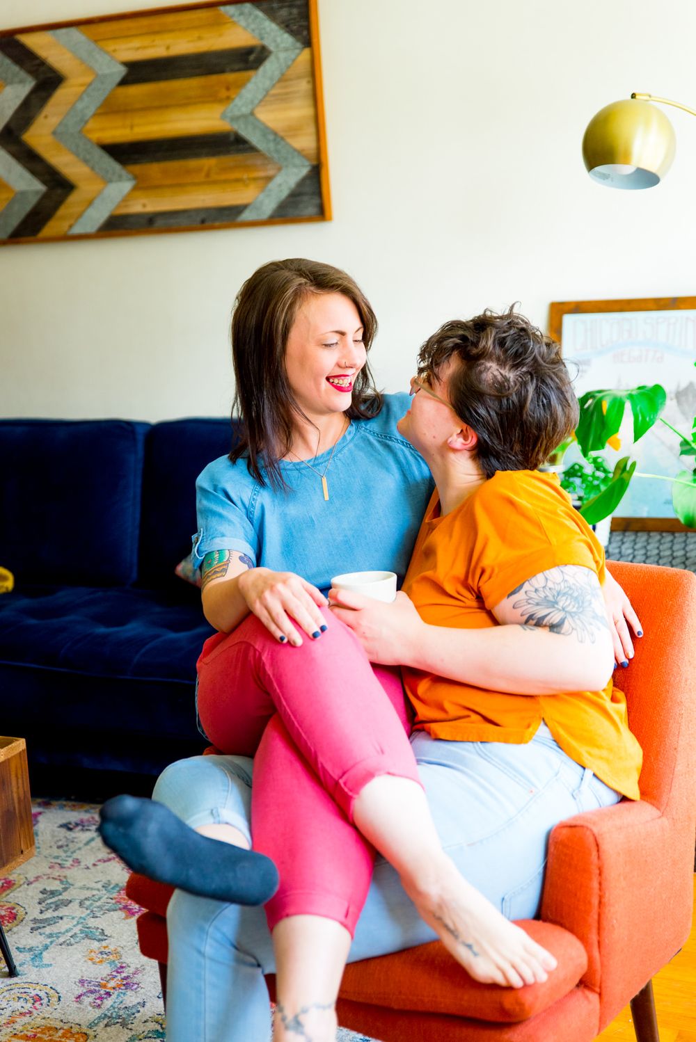 young woman in blue shirt and pink pants sitting on lap of wife in orange shirt with short hair on orange chair