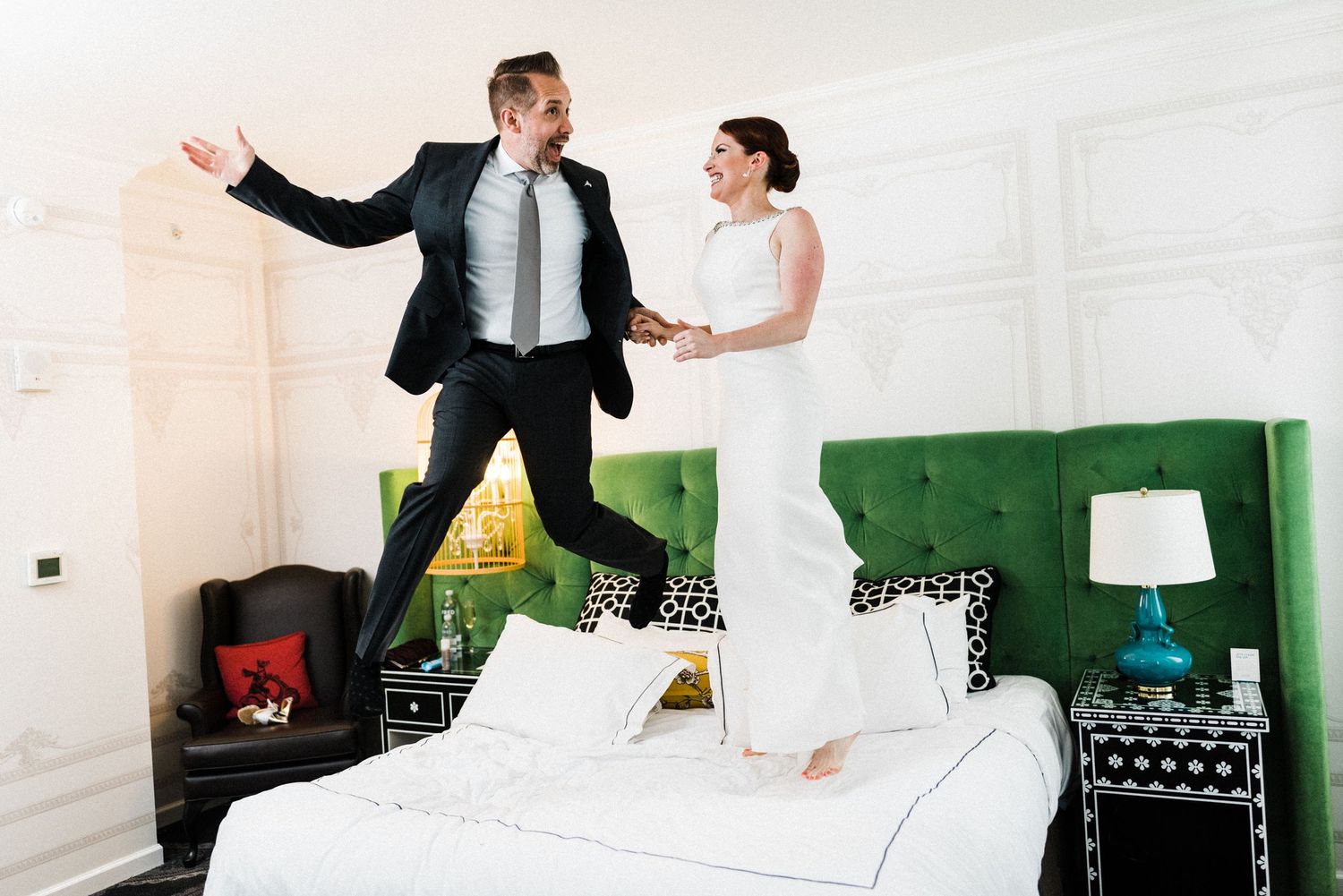 Playful couple jumping on hotel bed in formal attire.