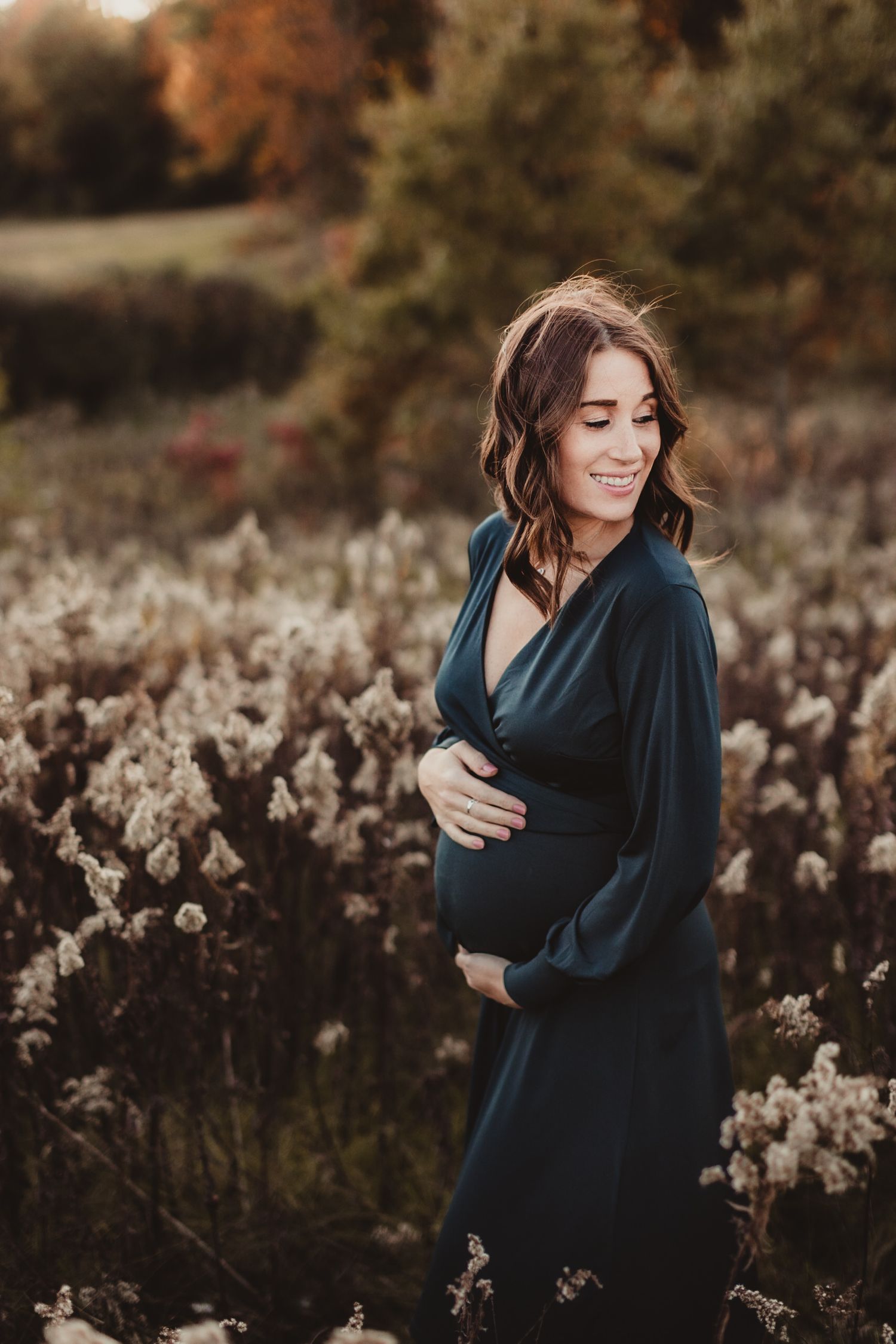 A woman cradles her pregnant belly in a fall season meadow. Photo by maternity photographer, Elizabeth Pachniak.