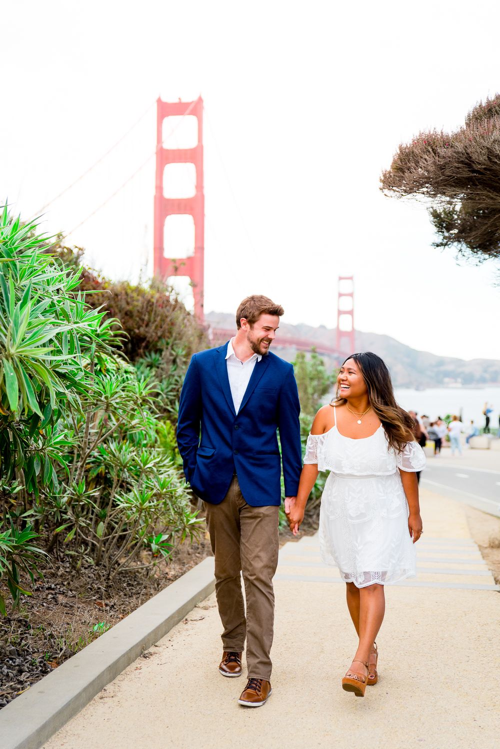 man in navy suit and woman in white dress holding hands laughing at Golden Gate Park engagement pictures